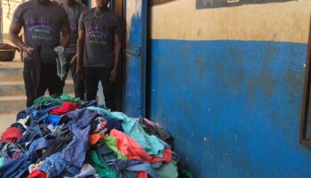 Three men stand beside a large pile of colorful clothes in front of a blue and yellow building.